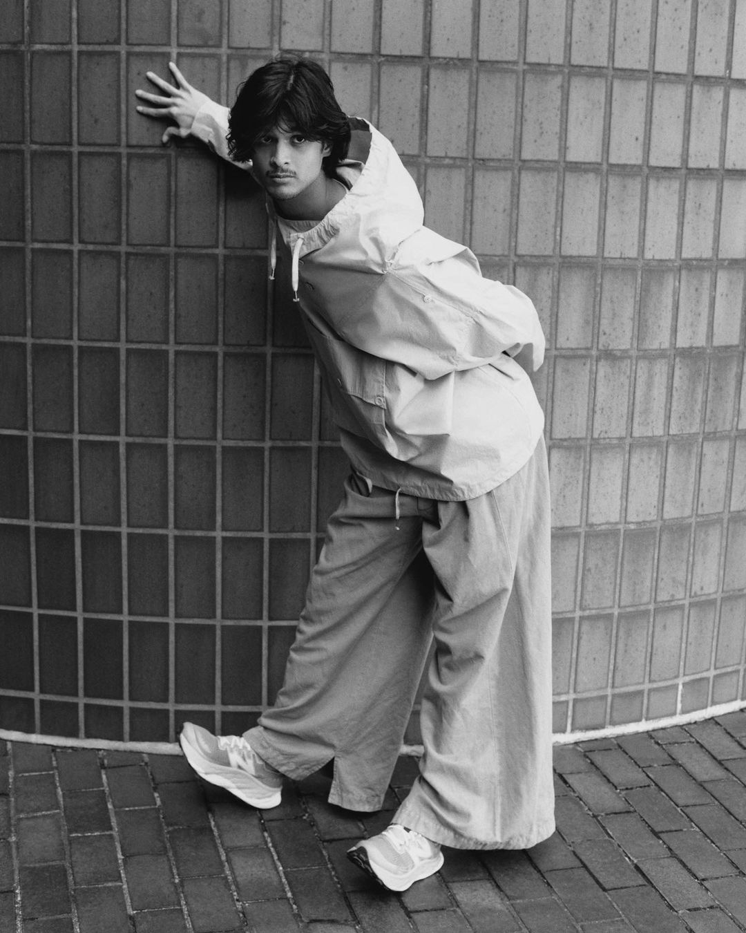 Monochrome portrait of a young man leaning against a curved tiled wall wearing a windbreaker and wide pants.