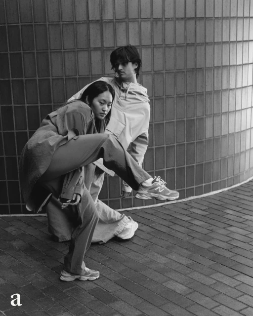 Two people pose in front of textured wall: Asian woman in oversized tan suit balances on one leg, while the blurry male figure poses behind her.