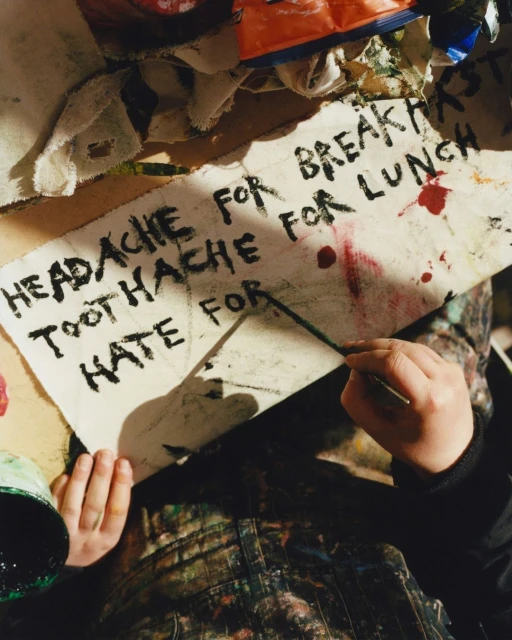 Child artist drawing on a canvas with black marker, wearing paint-covered overalls, in an indoor art studio setting. 