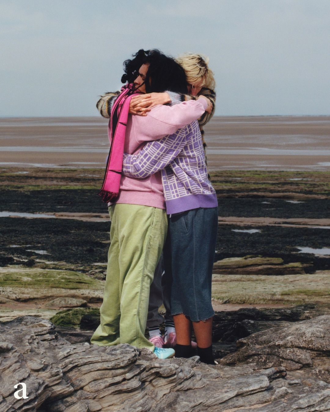 Three people embracing on a rocky beach. The figures, facing away from the camera, wear casual sweaters and trousers against a coastal backdrop.