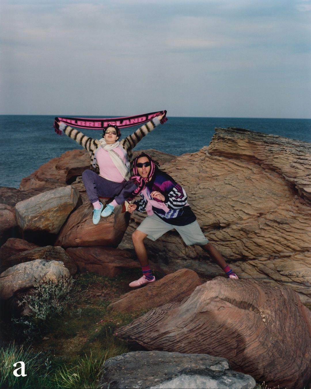 Two people pose on rocks overlooking the ocean. One holds a pink scarf. Both wear sunglasses and colorful vintage clothing.