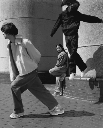 Black and white shot of three young adults in casual clothing, posing in front of a textured concrete wall on a brick surface outdoors.