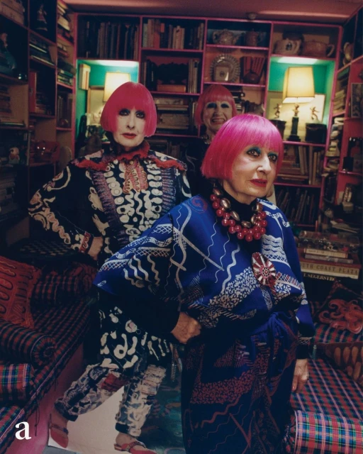 Three stylish women with pink bob haircuts pose in an eclectic room with patterned wallpaper, carpets, and overflowing bookshelves in the background.