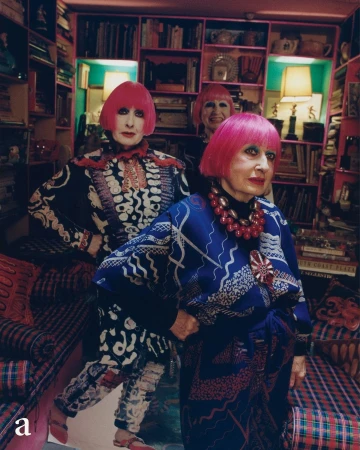 Three stylish women with pink bob haircuts pose in an eclectic room with patterned wallpaper, carpets, and overflowing bookshelves in the background.