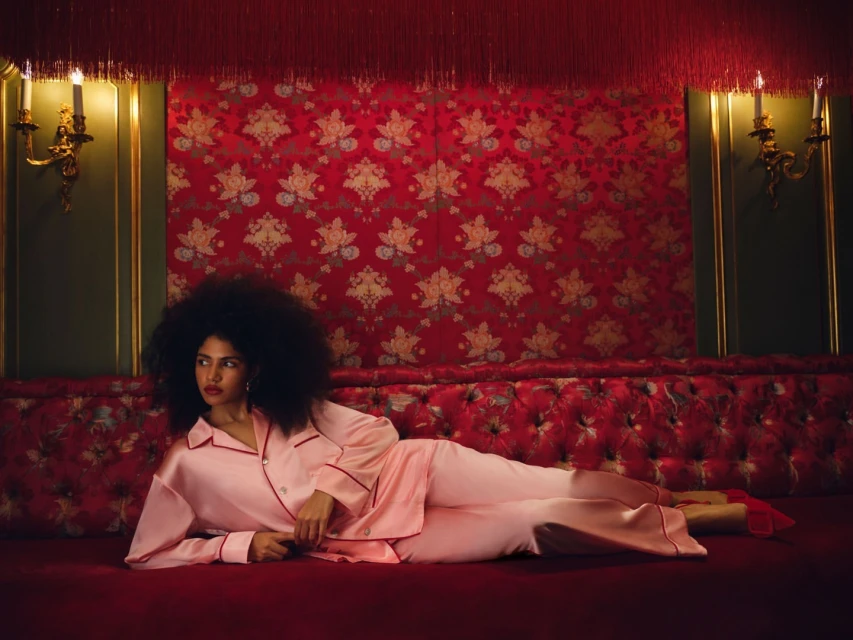 Woman with afro lying on red patterned chaise wearing pink silk pajamas and red heels against a red floral wallpapered wall.