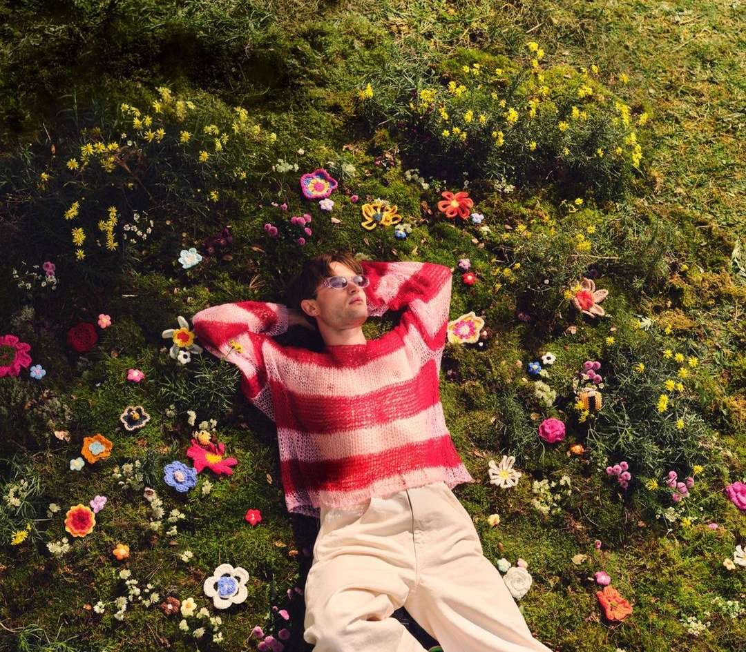 An adult male lies on a mossy surface covered in handmade yarn flowers, wearing glasses and a striped red and white sweater. 