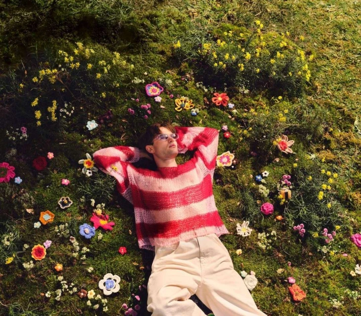 An adult male lies on a mossy surface covered in handmade yarn flowers, wearing glasses and a striped red and white sweater. 