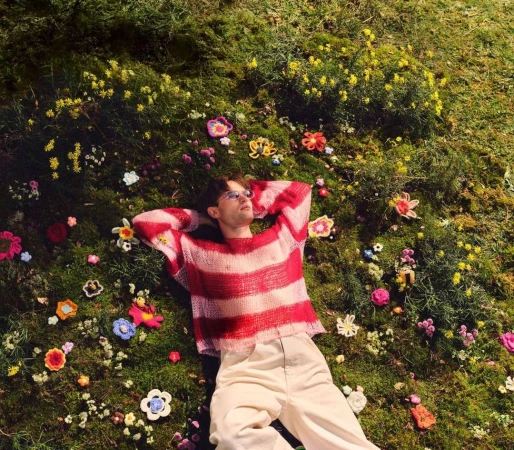 An adult male lies on a mossy surface covered in handmade yarn flowers, wearing glasses and a striped red and white sweater. 