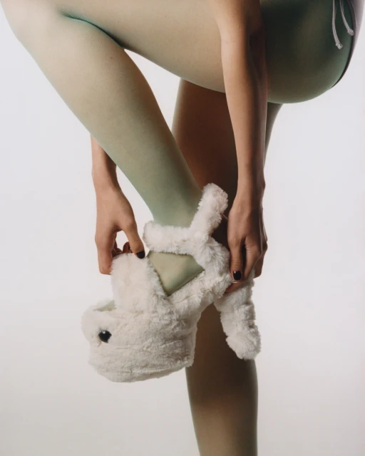 Close-up studio shot of an adult female in light green tights, adjusting a plush white novelty bunny slipper/shoe on her right foot.