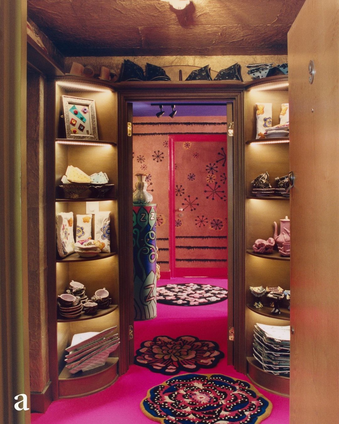 Eclectic interior with pink floor, rugs, and shelving displaying dishware, pottery, and decor in a room with a doorway and patterned walls.