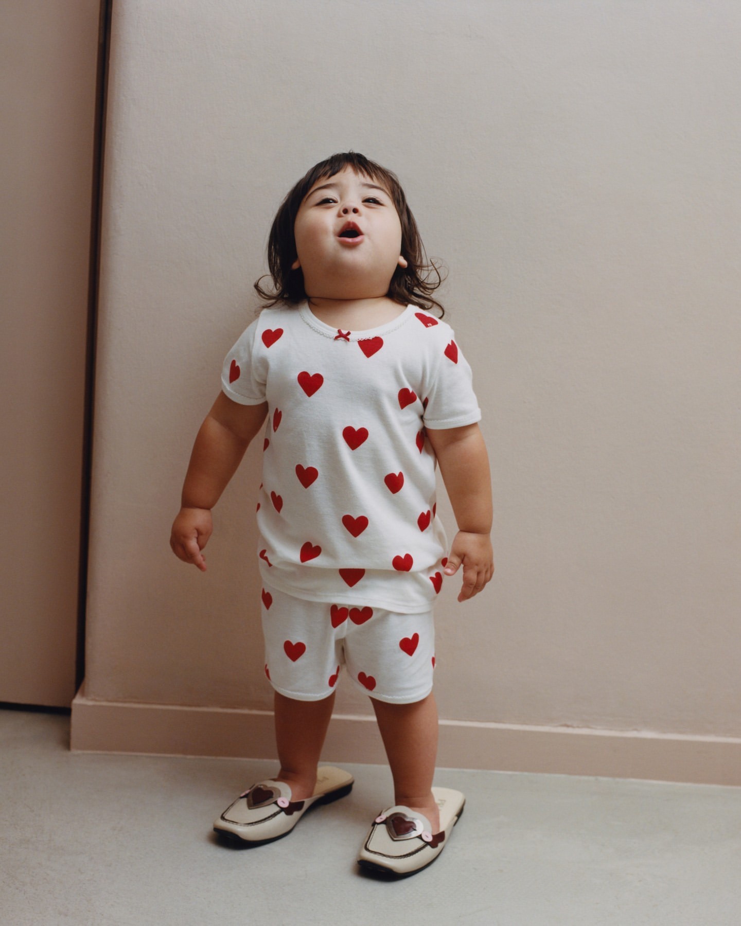 Full shot of a child in matching white pajamas with red hearts, standing indoors looking up while wearing adult-sized loafers.