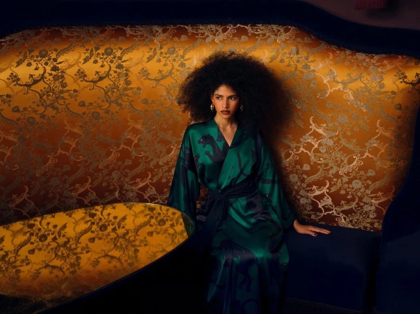 Elegant woman with large afro wearing a green and black patterned robe, seated on a blue couch with gold patterned backdrop, indoors.