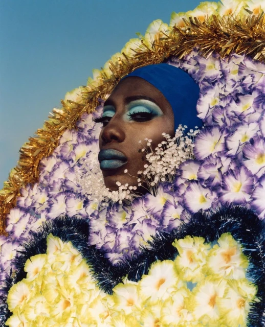 Portrait of a non-binary Black person in bright blue head wrap and blue lipstick, framed by a large floral decoration with gold tinsel.