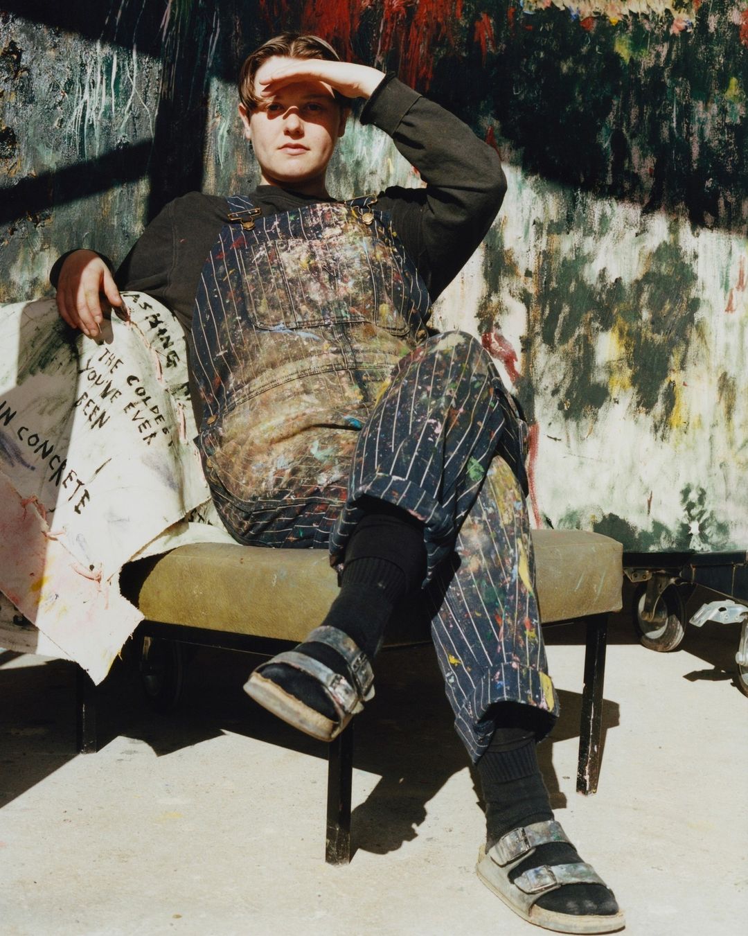 Adult male artist sits on chair in paint-splattered overalls, casting shade over his face in a paint-splattered room.