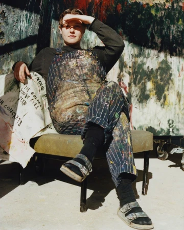 Adult male artist sits on chair in paint-splattered overalls, casting shade over his face in a paint-splattered room.