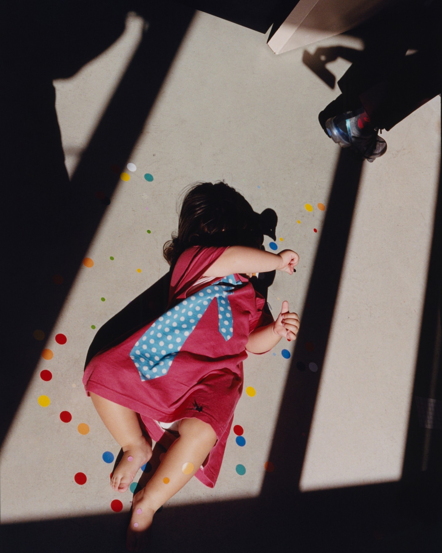 Overhead shot of a toddler lying on a gray surface surrounded by colorful confetti, wearing a red dress and a blue tie with white dots.
