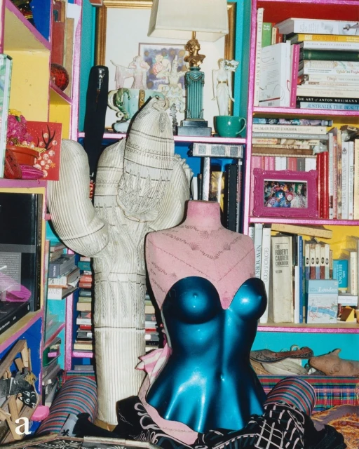 Eclectic interior featuring a pink and blue mannequin torso, a cardboard cactus, and a colorful bookshelf filled with books and decor.