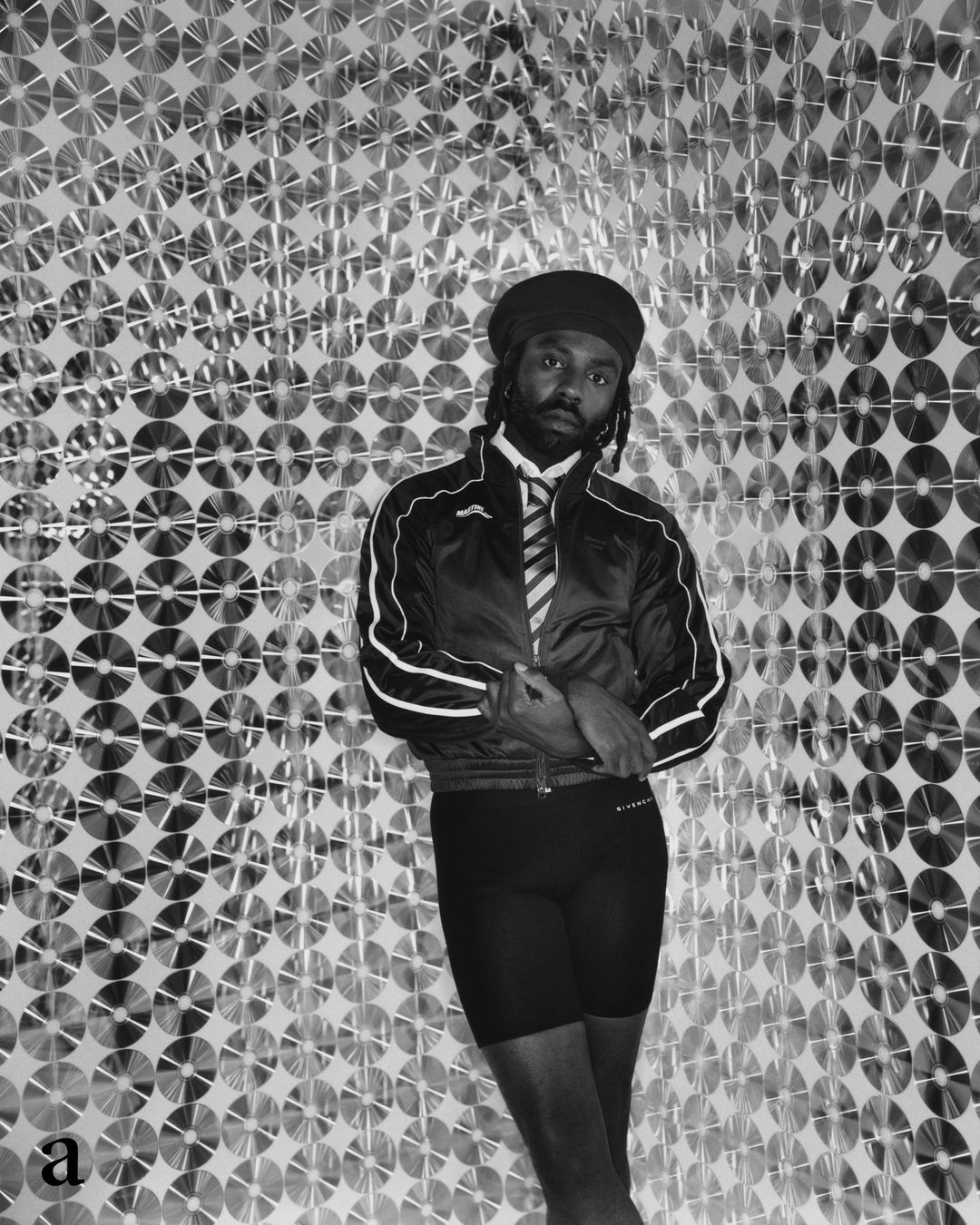 Man wearing a track jacket and cap posing against a shimmering wall of compact discs in a portrait by Joyce Ng.