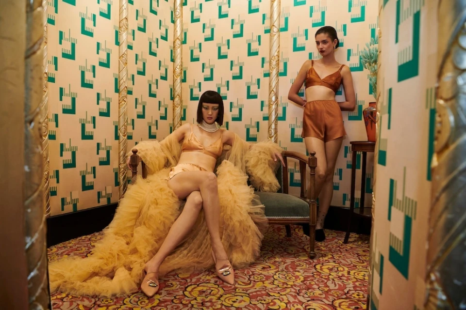 Two women model lingerie in a vintage-style room with patterned wallpaper and ornate decor. One sits in a chair wearing a yellow robe.