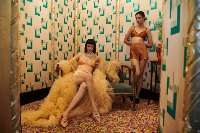 Two women model lingerie in a vintage-style room with patterned wallpaper and ornate decor. One sits in a chair wearing a yellow robe.