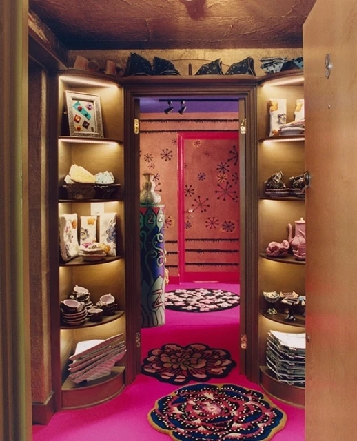 Eclectic interior view of a hallway with shelves displaying dishware and decorative items, leading to a pink room with round rugs.