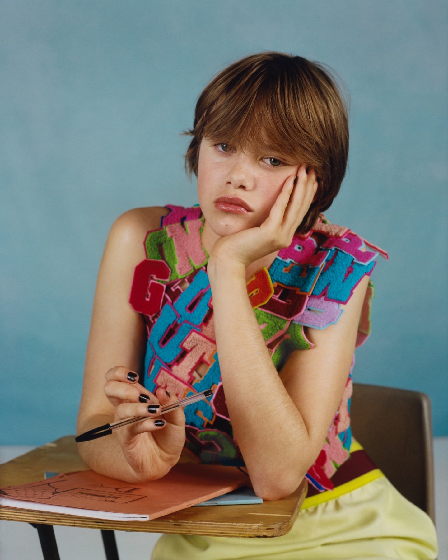 Teen girl in colorful top sits at a school desk, holding a pen with a muted blue background, creating an editorial fashion image.