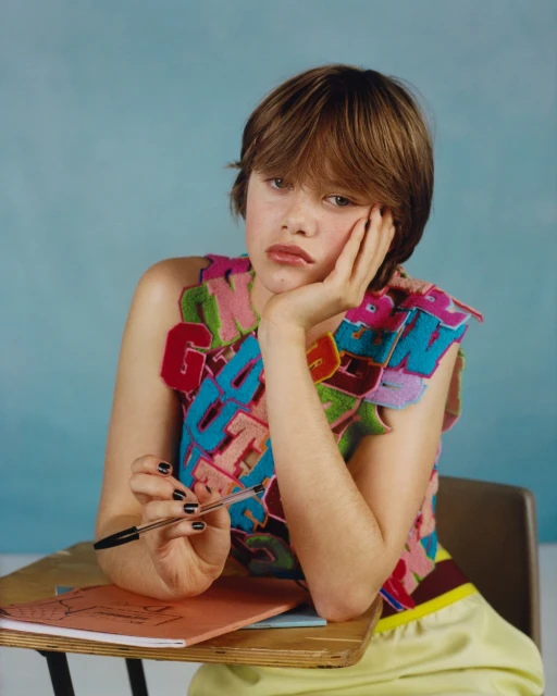 Teen girl in colorful top sits at a school desk, holding a pen with a muted blue background, creating an editorial fashion image.