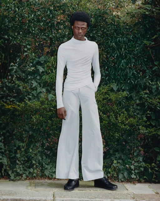 A slender adult male with dark complexion poses outdoors against a green foliage backdrop, wearing an all-white long-sleeved outfit with black shoes.