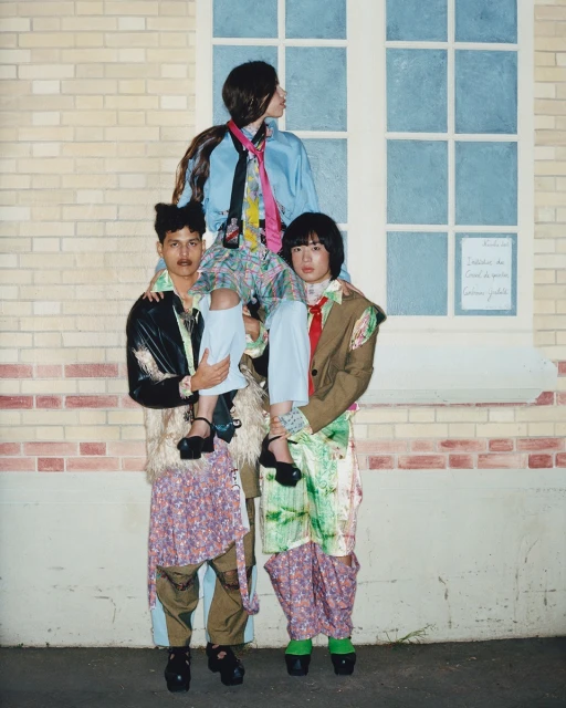 Three models stacked vertically in an outdoor editorial fashion shot. Two models support the third, posing in front of a brick building.