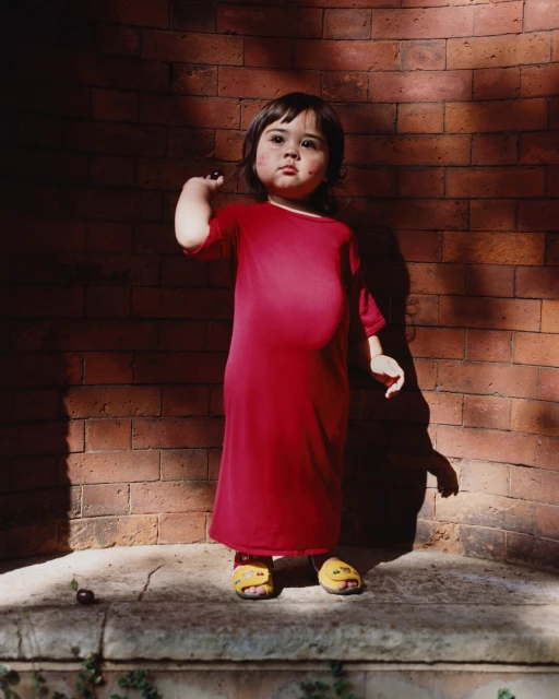 A young child with dark hair stands on a stone ledge in front of a brick wall, wearing a red dress and yellow sandals, holding a dark fruit.