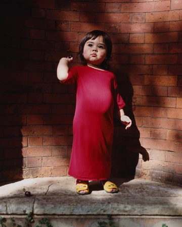 A young child with dark hair stands on a stone ledge in front of a brick wall, wearing a red dress and yellow sandals, holding a dark fruit.