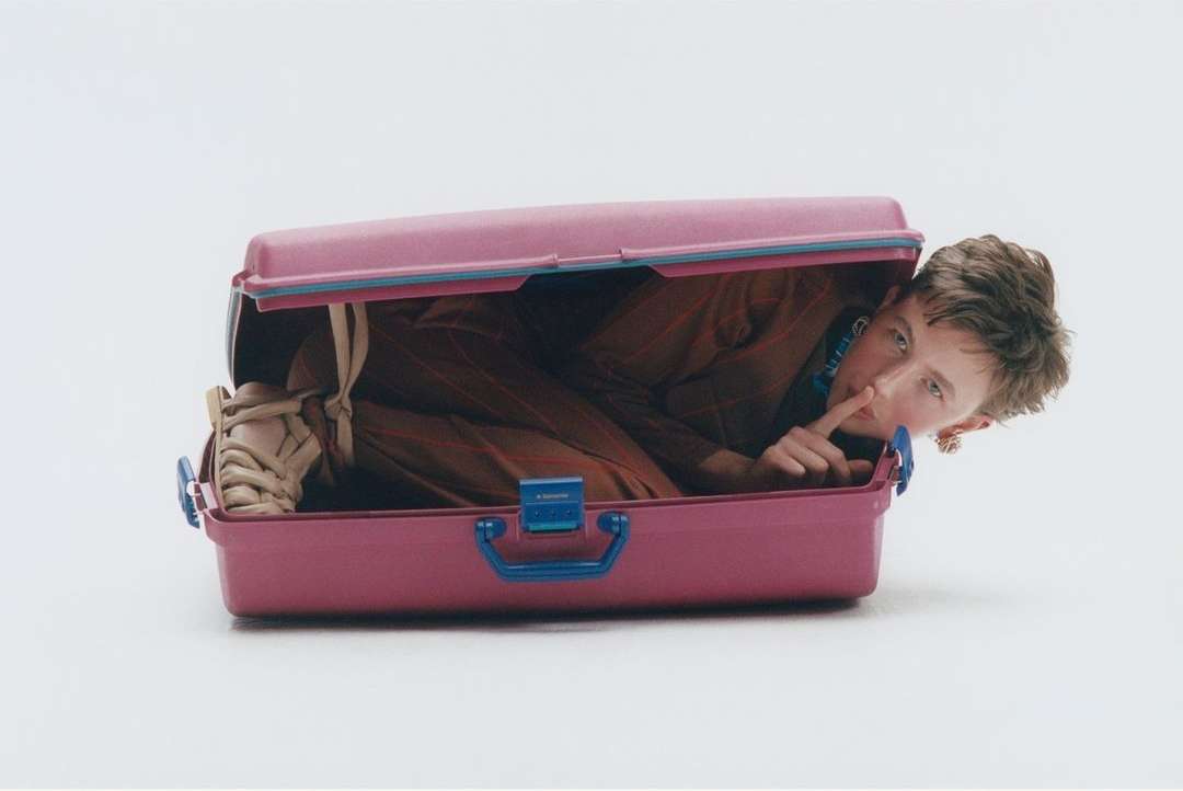 Young adult inside a pink suitcase with beige shoes sticking out, holding a finger to lips for silence, on a white background.
