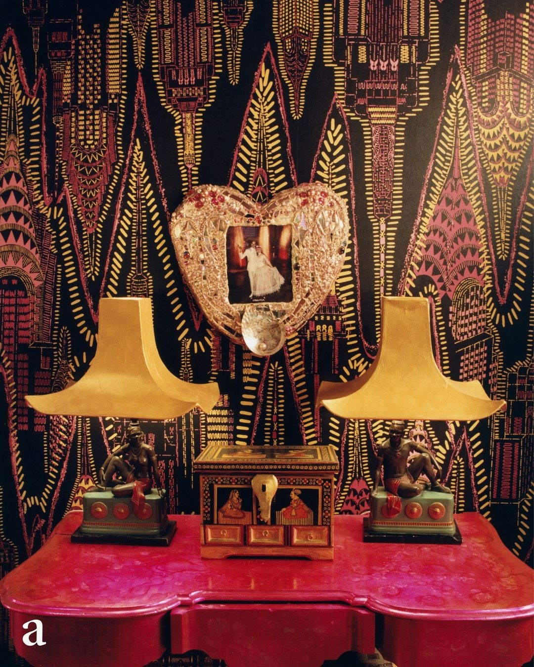 Interior still life features two gold lamps flanking an ornate box on a red table, set against a patterned wallpaper backdrop.