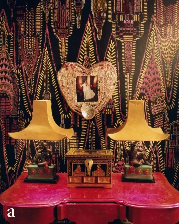 Interior still life features two gold lamps flanking an ornate box on a red table, set against a patterned wallpaper backdrop.