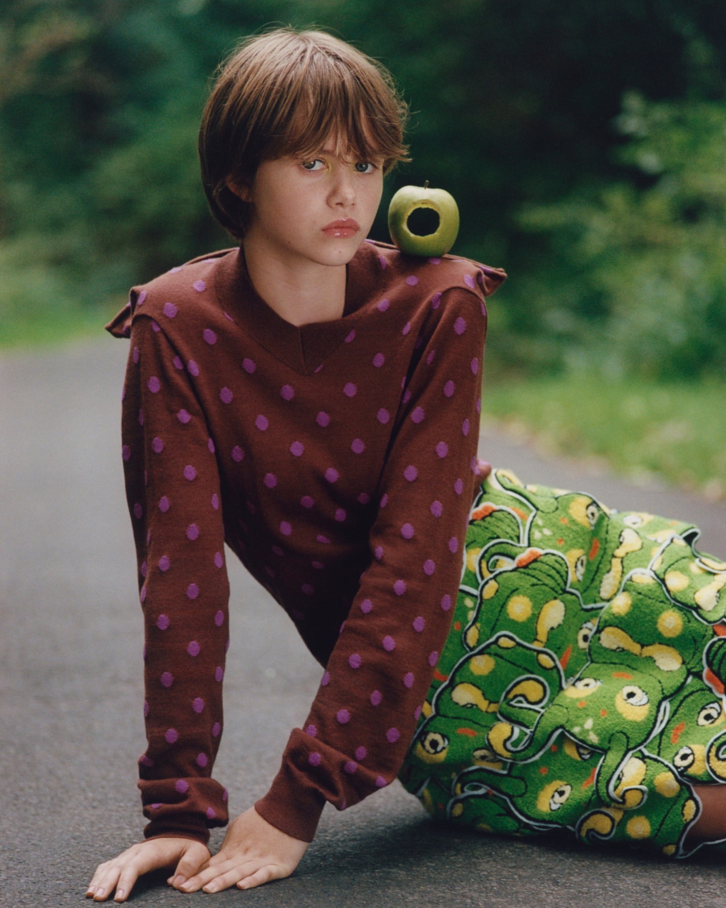 A young woman with an apple on her shoulder poses on a road in a brown dotted sweater and patterned skirt in a natural outdoor setting.