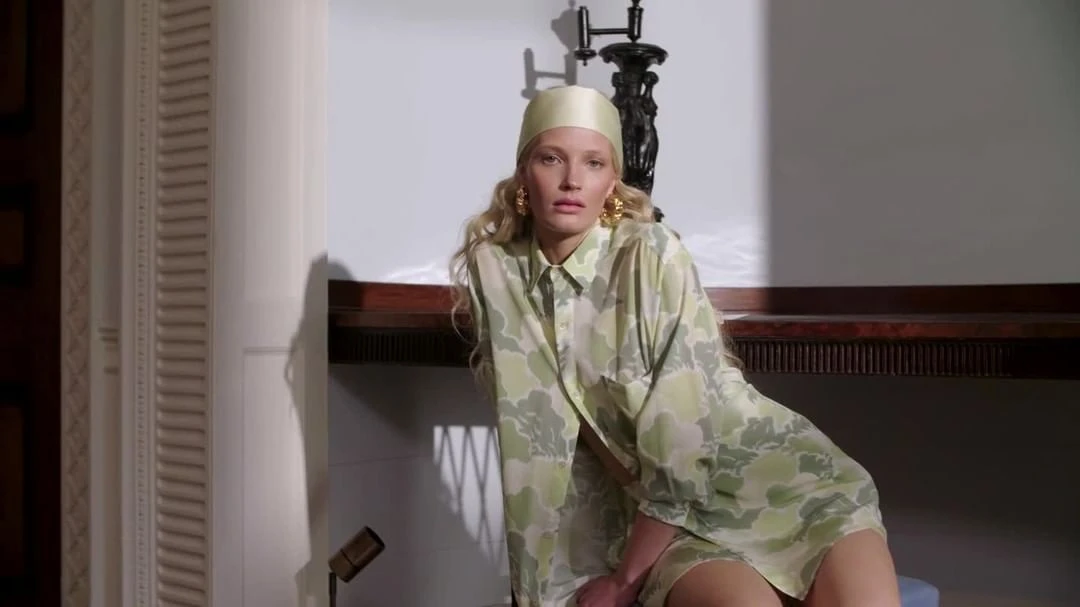 Blonde woman in green patterned shirt and cream-colored headscarf sits indoors near dark wood table and white decorated wall.
