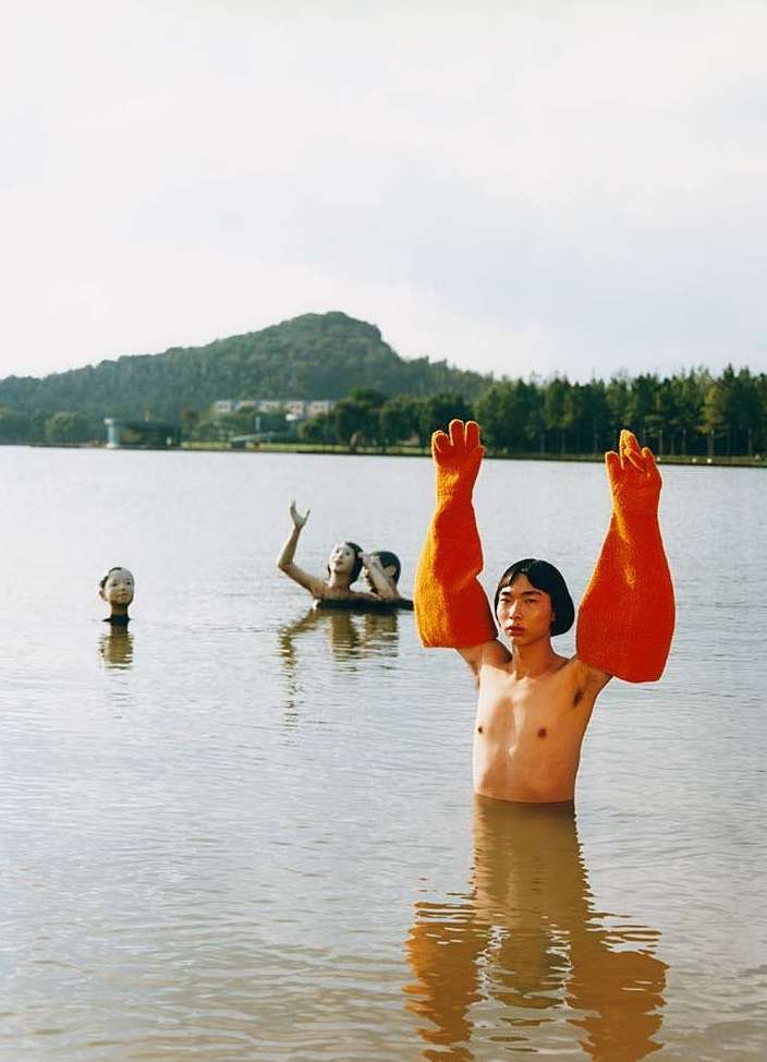 Artistic photograph in a lake featuring a man with orange gloves and partial figures with their arms raised against a green landscape backdrop.