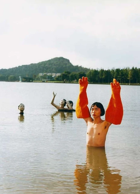 Artistic photograph in a lake featuring a man with orange gloves and partial figures with their arms raised against a green landscape backdrop.