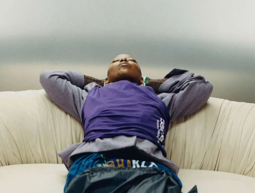 Person lounges indoors with eyes closed, wearing a purple shirt and grey jacket, hands behind head on a cream-colored couch.
