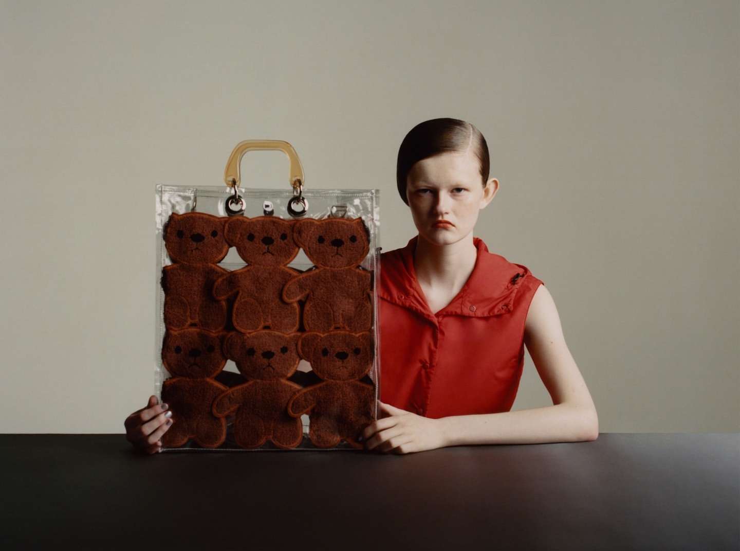 Fashion shot of a young adult woman with a red top, holding a clear tote bag filled with brown teddy bear toys in a studio setting.