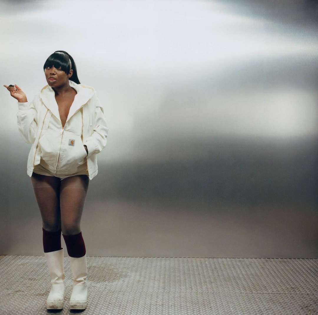 Stylish woman with a ponytail poses in a beige jacket, white shorts, high socks, and platform boots, standing against a metallic background.