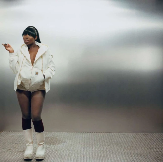 Stylish woman with a ponytail poses in a beige jacket, white shorts, high socks, and platform boots, standing against a metallic background.