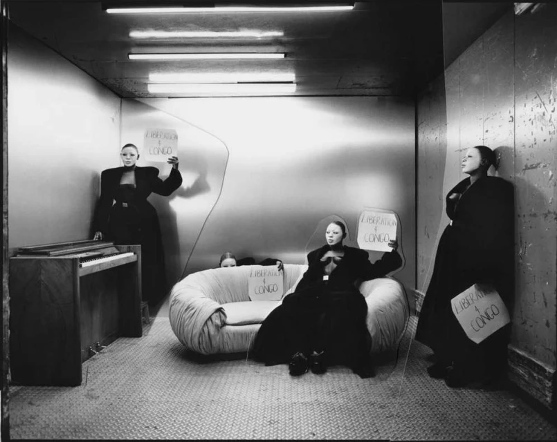 Monochrome studio fashion shot of four women in dark clothing with large shoulder pads holding signs in an enclosed, spare space.