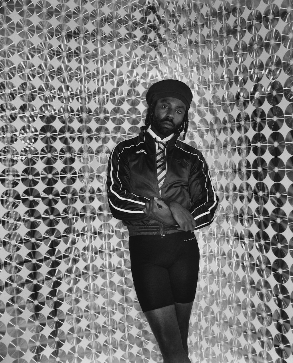 Portrait of African-American man in jacket, tie, and shorts posing indoors against a wall covered in rows of CDs.