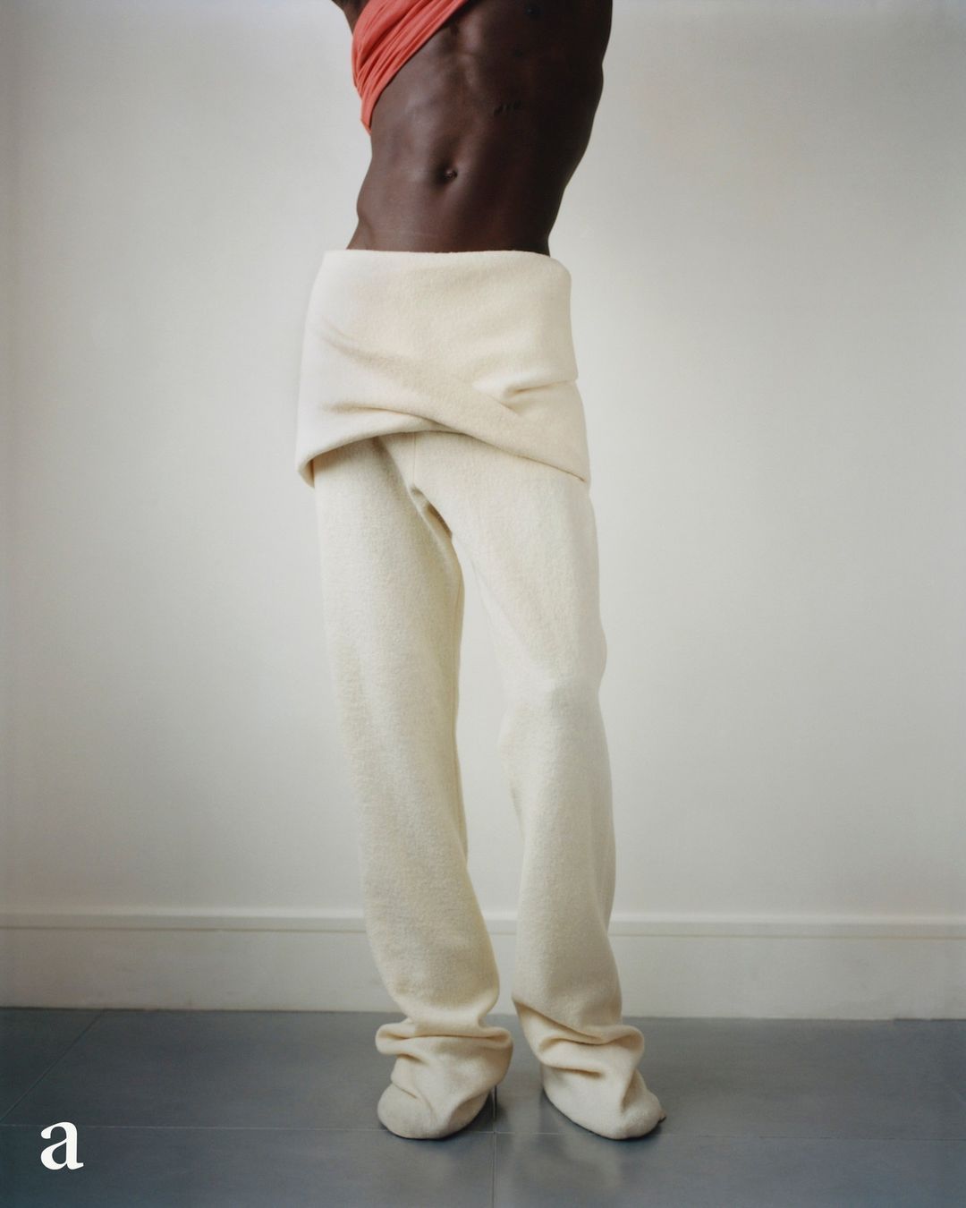 Dark-skinned person in off-white pants and coral top stands in front of a white wall with gray flooring, showcasing avant-garde fashion.