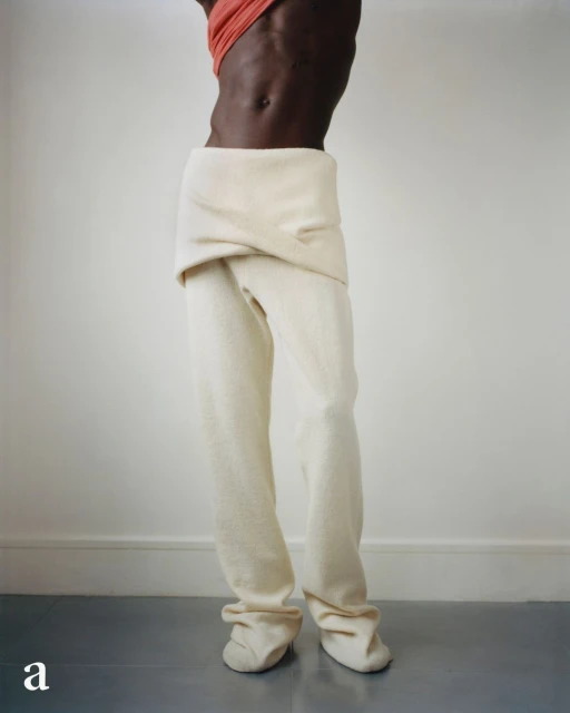 Dark-skinned person in off-white pants and coral top stands in front of a white wall with gray flooring, showcasing avant-garde fashion.