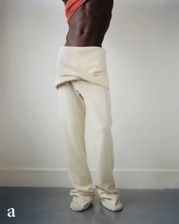 Dark-skinned person in off-white pants and coral top stands in front of a white wall with gray flooring, showcasing avant-garde fashion.