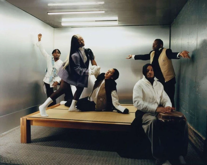 Group of five people in a bare room, posing on and around a low platform bed. Two women and three men, diverse attire and expressions.