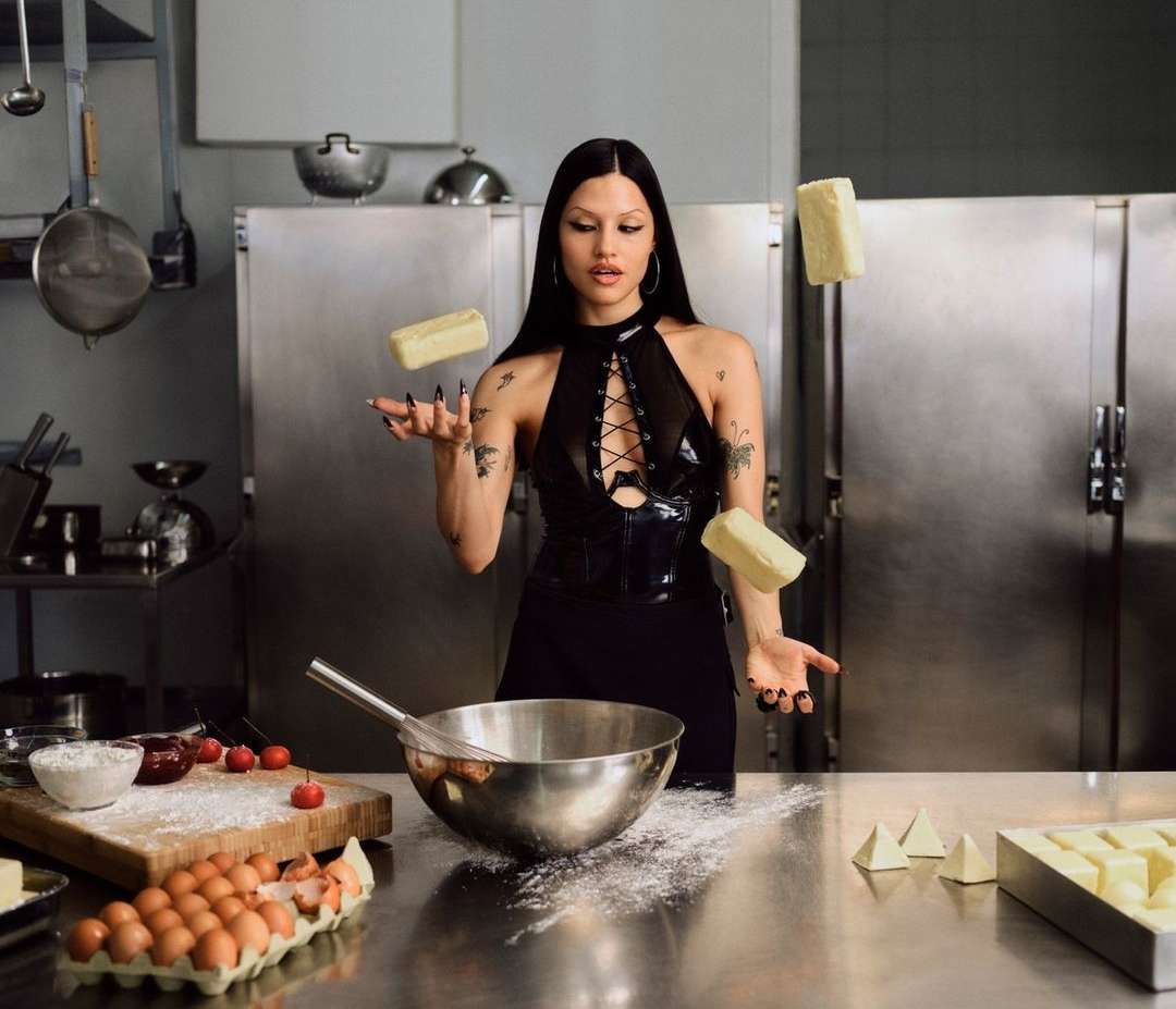 A woman in a black dress juggles blocks of butter in a kitchen, surrounded by baking ingredients and tools on a stainless steel counter.