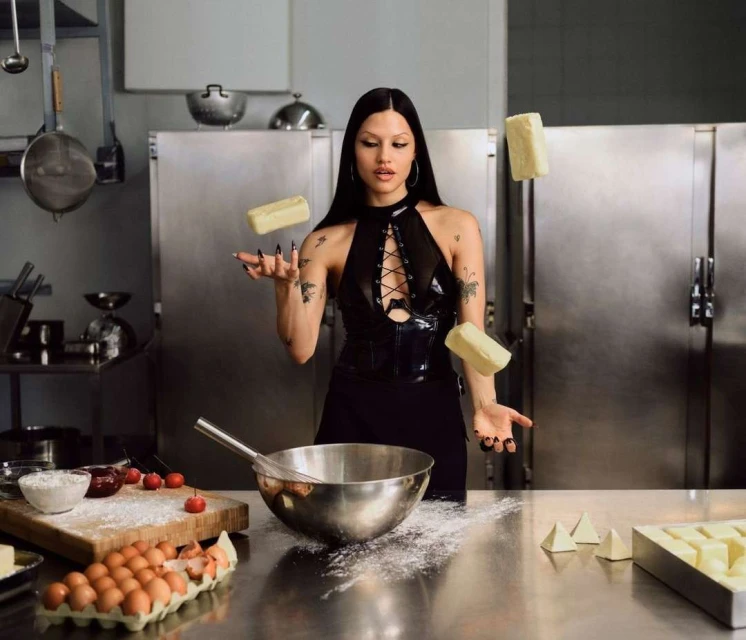A woman in a black dress juggles blocks of butter in a kitchen, surrounded by baking ingredients and tools on a stainless steel counter.